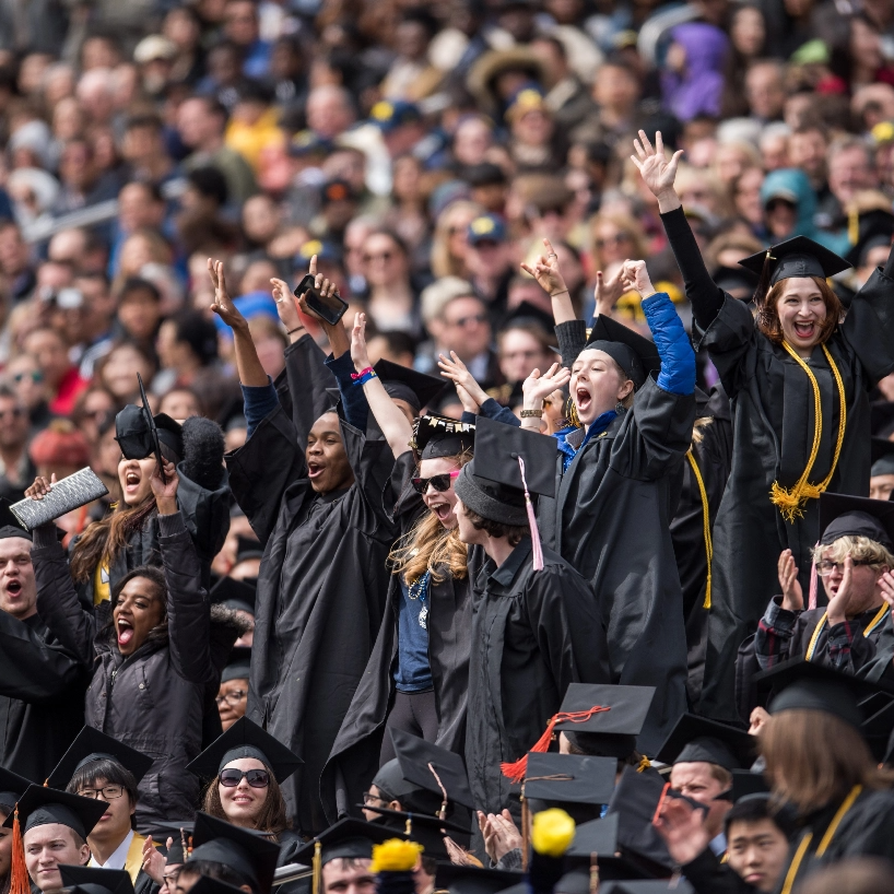A crowd of Michigan students wearing caps and gowns cheering at commencement