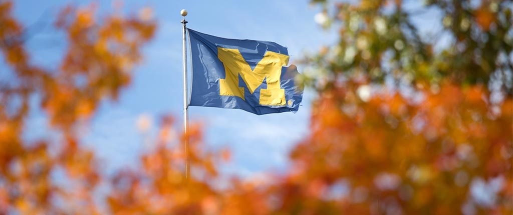 The block M flag is spotted beyond some fiery-colored trees in autumn.